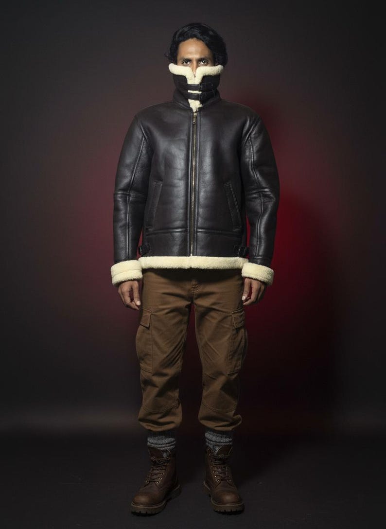 May include: A black leather jacket with a cream-colored shearling lining and trim. The jacket has a zip-up front and two pockets. The person is wearing brown cargo pants and brown boots. The background is a dark red.
