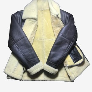 May include: A dark brown leather jacket with a cream-colored shearling lining and collar. The jacket has a classic design with a zip closure and a pocket on the front. The sleeves are long and have a shearling trim at the cuffs. The jacket is laid flat.