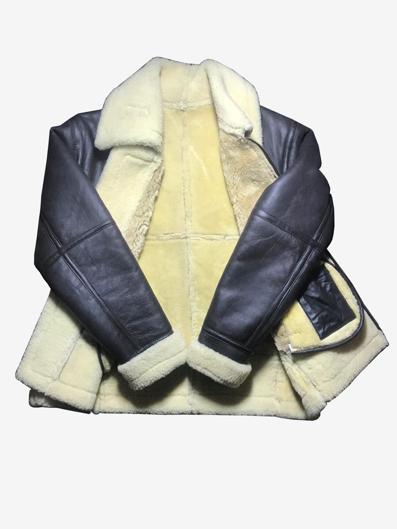 May include: A dark brown leather jacket with a cream-colored shearling lining and collar. The jacket has a classic design with a zip closure and a pocket on the front. The sleeves are long and have a shearling trim at the cuffs. The jacket is laid flat.