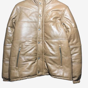 K&ouml;nnte beinhalten: Hellbraune Leder-Steppjacke mit hohem Kragen und Druckknopf- und Rei&szlig;verschl&uuml;ssen. Die Jacke hat ein gestepptes Design und dekorative N&auml;hte an den Schultern. Zwei Rei&szlig;verschlusstaschen sind auf der Vorderseite sichtbar.