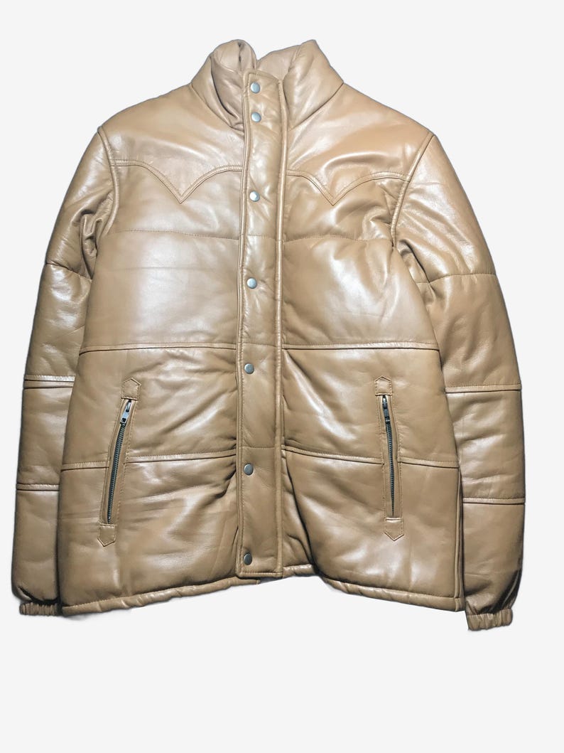 K&ouml;nnte beinhalten: Hellbraune Leder-Steppjacke mit hohem Kragen und Druckknopf- und Rei&szlig;verschl&uuml;ssen. Die Jacke hat ein gestepptes Design und dekorative N&auml;hte an den Schultern. Zwei Rei&szlig;verschlusstaschen sind auf der Vorderseite sichtbar.