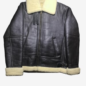 May include: A dark brown leather jacket with a cream-colored shearling collar and lining. The jacket features a front zipper, two front pockets, and a buckle at the waist. The cuffs are also lined with cream-colored shearling.