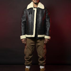 May include: Black leather jacket with a cream-colored shearling collar and trim. The jacket is worn with brown cargo pants and tan work boots. The background is a dark maroon color.