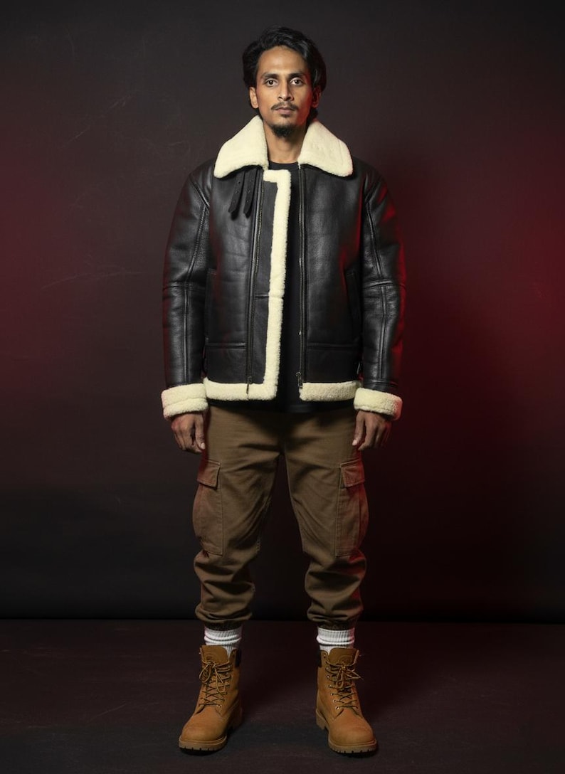 May include: Black leather jacket with a cream-colored shearling collar and trim. The jacket is worn with brown cargo pants and tan work boots. The background is a dark maroon color.