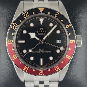 May include: A stainless steel wristwatch with a black dial and a red and black bezel. The watch features a date window at the 3 o'clock position and a silver-tone bracelet. The watch face has the text "TUDOR GENEVE GMT MASTER CHRONOMETER SWISS MADE".