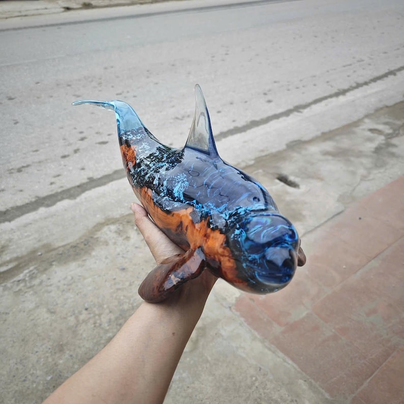 Może przedstawiać: Rzeźba delfina wykonana z połączenia polerowanego naturalnego drewna i p&oacute;łprzezroczystej niebieskiej żywicy. Przedmiot ma gładkie, błyszczące wykończenie, przez kt&oacute;re widać słoje drewna, prezentując smukłą, zakrzywioną formę wodnego zwierzęcia.