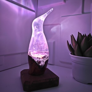 Könnte beinhalten: Dekorative Lampe mit klarer, geschwungener Oberseite und Holzsockel. Das Innere der Lampe zeigt eine Landschaftsszene mit lila und rosa Farbtönen. Die Lampe ist beleuchtet und strahlt sanft.