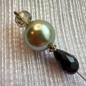 Può includere: Spilla decorativa con una grande perla verde perlescente, una perla nera sfaccettata a forma di goccia e una piccola perla grigia sfaccettata. La spilla ha dettagli color oro e un sottile spillo color argento.