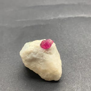 以下が含まれることがあります： 上部に鮮やかな赤いルビーの結晶が埋め込まれた、小さく、ざらざらにカットされたオフホワイトの石。ルビーは深く飽和した赤色です。石は濃い灰色の背景に配置されています。