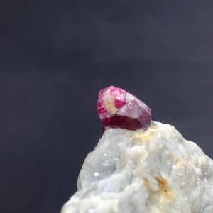 May include: A close-up of a rough, natural ruby crystal with a deep red hue, accented by lighter pink and white areas. The ruby sits atop a light-coloured, textured rock formation, set against a dark grey background.