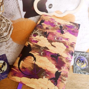 May include: A book cover with a fantasy theme, featuring dragons, clouds, and celestial elements in shades of purple, brown, and gold. The design includes silhouettes of dragons and suns.