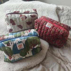 Peut inclure: Trois trousses de maquillage matelassées avec différents motifs. L'une présente un fond blanc avec des motifs de feuilles marron, une autre un motif floral rouge et la troisième un motif de montagne et d'arbres bleu et vert. Chaque trousse a une fermeture éclair.