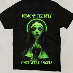 Op de afbeelding: Zwart t-shirt met een afbeelding van een non die de middelvinger opsteekt, een groene halo en de tekst "DEMONY TEŽ BLY" en "ONCE WERE ANGELS". Het ontwerp heeft een gotisch en rebels thema.