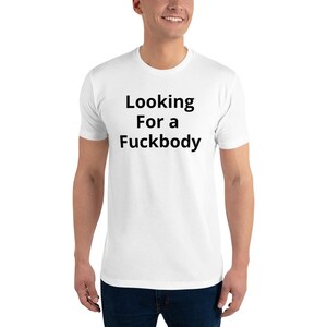 Puede incluir: Camiseta blanca con el texto "Looking For a Fuckbody" en negro, centrado en el pecho. La camiseta es de manga corta y parece estar hecha de un material suave. La persona que lleva la camiseta está sonriendo.