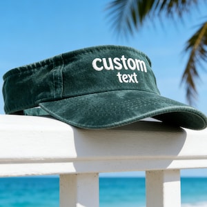 May include: A dark green visor with the words "custom text" embroidered in white. The visor is resting on a white railing with a beach and ocean in the background. A palm tree is visible in the background.