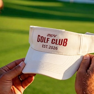 May include: An off-white visor featuring the embroidered text SHOW GOLF CLUB in a bold, dark red font, with the smaller text EST. 2026 positioned directly underneath. The visor has a structured front panel and a curved brim with stitched detailing.