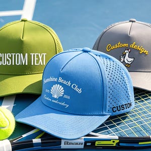 May include: Three baseball caps in olive green, bright blue, and light grey. The green cap features the text CUSTOM TEXT. The blue cap displays Sunshine Beach Club with a shell graphic and the word CUSTOM. The grey cap shows a duck graphic with the text Custom design.