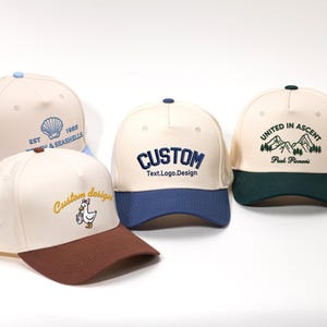 May include: Several baseball caps in cream, brown, blue, and green. Each cap has embroidered designs, including a seashell, the word "CUSTOM", a mountain scene, and a duck with a bucket. The caps have curved brims.