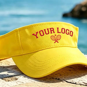 May include: A bright yellow sun visor featuring the red embroidered text "YOUR LOGO" arched over a graphic of two crossed tennis rackets and a small tennis ball. The visor has a curved brim with visible stitching and an adjustable back strap.