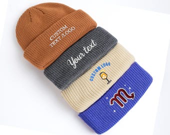 May include: A stack of five knit beanies in various colors: brown, gray, beige, and blue. Each beanie features embroidered text or a logo, including "CUSTOM TEXT/LOGO" and "Your text". The beanies are angled, showcasing their folded brims.