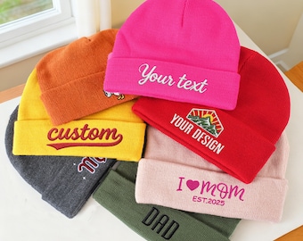 May include: A collection of colorful beanies in various shades, including pink, red, yellow, and green. Several beanies feature embroidered text such as "Your text", "Custom", "Your design", "I ❤️ mom", and "Dad".