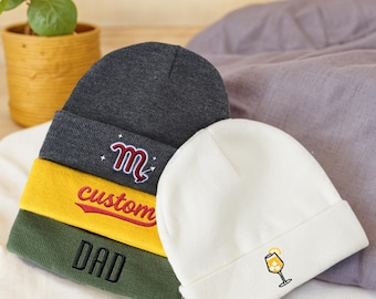 Op de afbeelding: Een verzameling mutsen in verschillende kleuren, waaronder grijs, geel, olijfgroen en wit. De mutsen hebben geborduurde ontwerpen en tekst, zoals een gestileerde "m", het woord "custom", "DAD" en een cocktailglas. De mutsen zijn omgeslagen aan de rand.