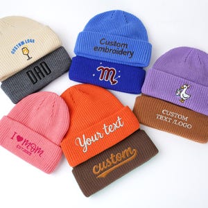 May include: A collection of colorful knit beanies with embroidered designs. The beanies are in various colors, including cream, pink, orange, blue, and purple. Each beanie features custom text or logos, such as "DAD," "I MOM," and "Your text."