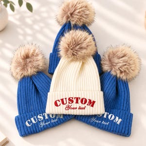 Puede incluir: Tres gorros de punto acanalado con grandes pompones de piel sintética. Dos gorros son azul real con texto bordado en blanco CUSTOM Your text, y uno central es color crema con texto bordado en rojo CUSTOM Your text.