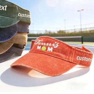 May include: An orange visor with the text "Tennis Mom" and a tennis ball graphic. The word "custom" is embroidered on the side. A stack of visors in various colours is visible in the background.