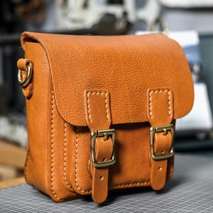 Op de afbeelding: Een kleine, rechthoekige, bruinleren tas met een klepsluiting en twee messing gespen. De tas heeft een schouderriem bevestiging en zichtbare witte stiksels. Het leer heeft een getextureerde uitstraling.