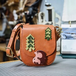 Op de afbeelding: Bruinleren crossbodytas met flap. De tas heeft groene en bruine vilten applicaties van bomen en een egel. De verstelbare riem en sluiting zijn bruin en goudkleurig. Een praktische tas voor dagelijks gebruik.