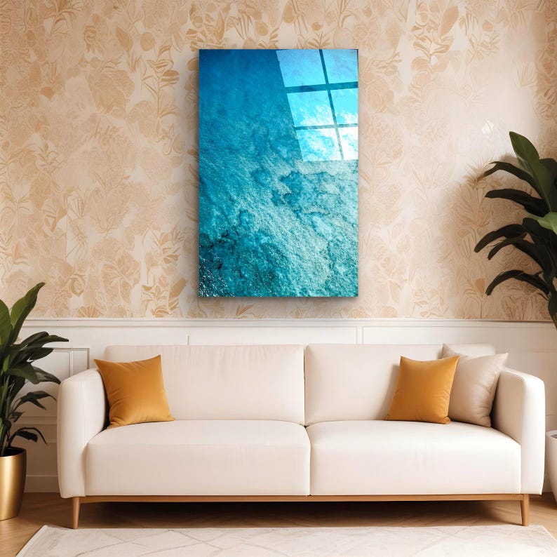 Puede incluir: Una obra de arte mural rectangular con un dise&ntilde;o azul y turquesa texturizado, que refleja la luz. La obra de arte cuelga sobre un sof&aacute; blanco con cojines naranjas, complementado con plantas en macetas. La habitaci&oacute;n tiene un papel pintado floral.