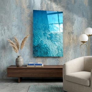 Puede incluir: Una pieza de arte mural rectangular con un vibrante dise&ntilde;o azul similar al oc&eacute;ano. La obra de arte est&aacute; montada en una pared texturizada de color azul claro. La imagen refleja la luz, y la escena incluye un mueble de madera, un jarr&oacute;n con plantas secas y una silla moderna.
