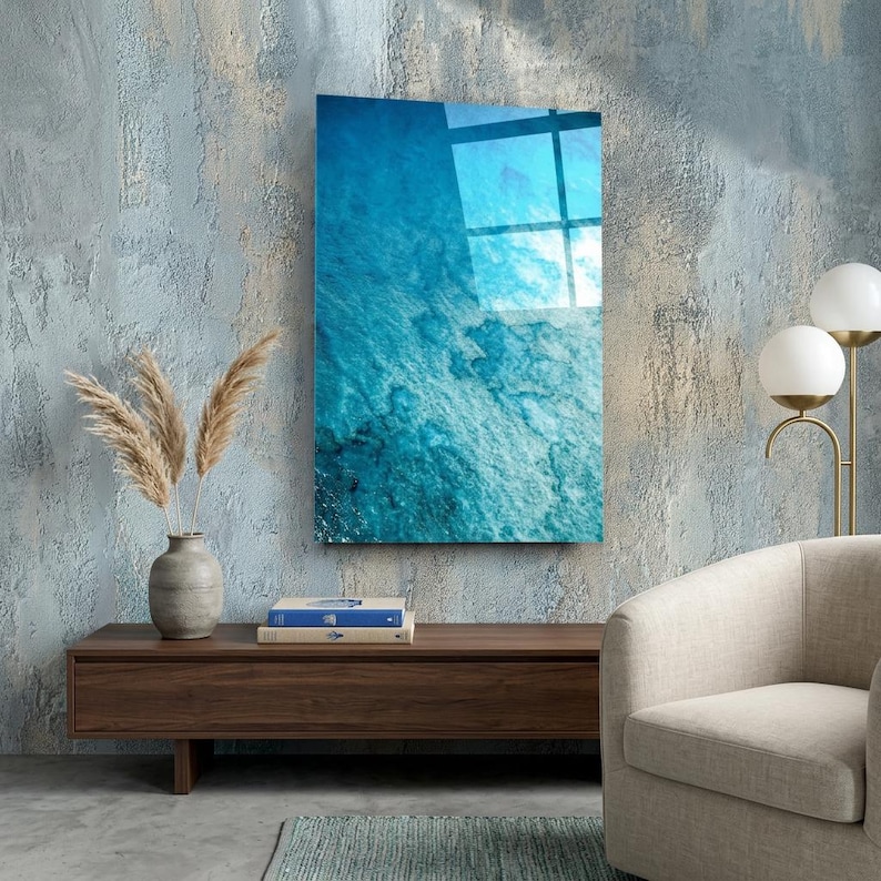 Puede incluir: Una pieza de arte mural rectangular con un vibrante dise&ntilde;o azul similar al oc&eacute;ano. La obra de arte est&aacute; montada en una pared texturizada de color azul claro. La imagen refleja la luz, y la escena incluye un mueble de madera, un jarr&oacute;n con plantas secas y una silla moderna.