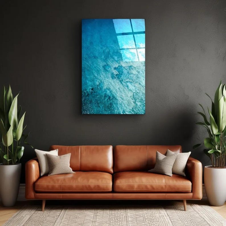 Puede incluir: Una obra de arte mural rectangular con un dise&ntilde;o abstracto azul y blanco, que se asemeja al agua o a una superficie texturizada. La obra de arte est&aacute; montada en una pared gris oscuro sobre un sof&aacute; de cuero marr&oacute;n con cojines. Plantas en macetas a ambos lados.