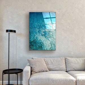 Puede incluir: Una obra de arte mural rectangular con un dise&ntilde;o abstracto texturizado en tonos azules y blancos. La obra de arte est&aacute; montada en una pared de color neutro sobre un sof&aacute; de color claro. La imagen refleja una ventana.