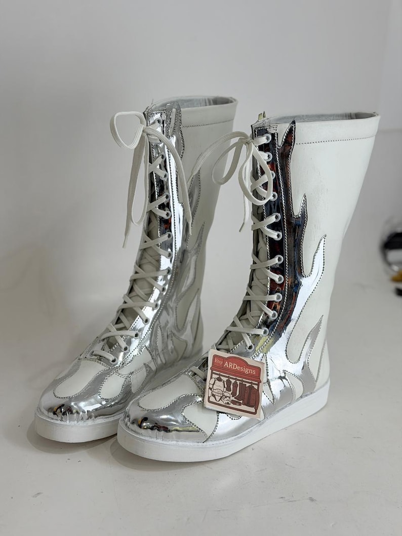 Puede incluir: Un par de botas altas blancas y plateadas con un dise&ntilde;o de llamas. Las botas tienen cordones blancos y una suela blanca. Las botas tienen una etiqueta que dice "ARDesigns".