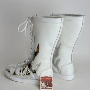 Puede incluir: Par de botas altas blancas con dise&ntilde;os de llamas plateadas y puntera plateada. Las botas tienen cordones blancos y cierre de cremallera. Una peque&ntilde;a etiqueta con el texto "ARDesigns" es visible.