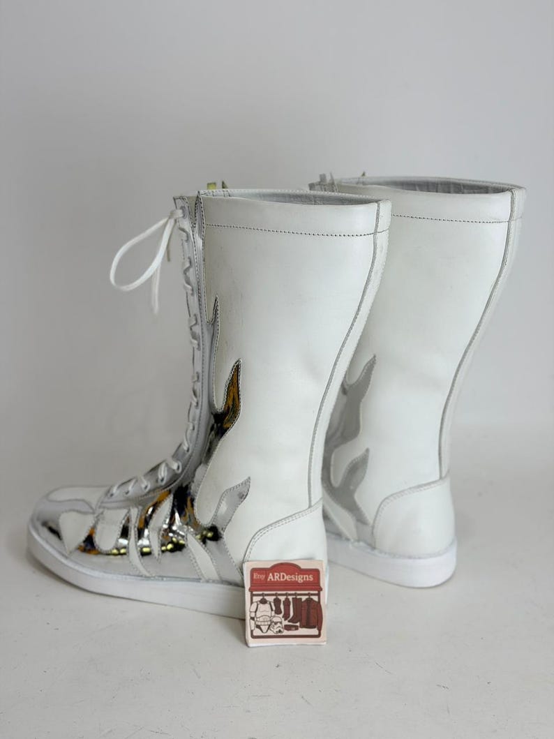Puede incluir: Par de botas altas blancas con dise&ntilde;os de llamas plateadas y puntera plateada. Las botas tienen cordones blancos y cierre de cremallera. Una peque&ntilde;a etiqueta con el texto "ARDesigns" es visible.
