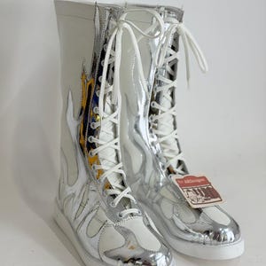 Puede incluir: Botas altas con un dise&ntilde;o de llamas blanco y plateado. Las botas tienen cordones blancos y una suela blanca. La parte superior de las botas es blanca, con detalles plateados en forma de llama. Una etiqueta est&aacute; adjunta.