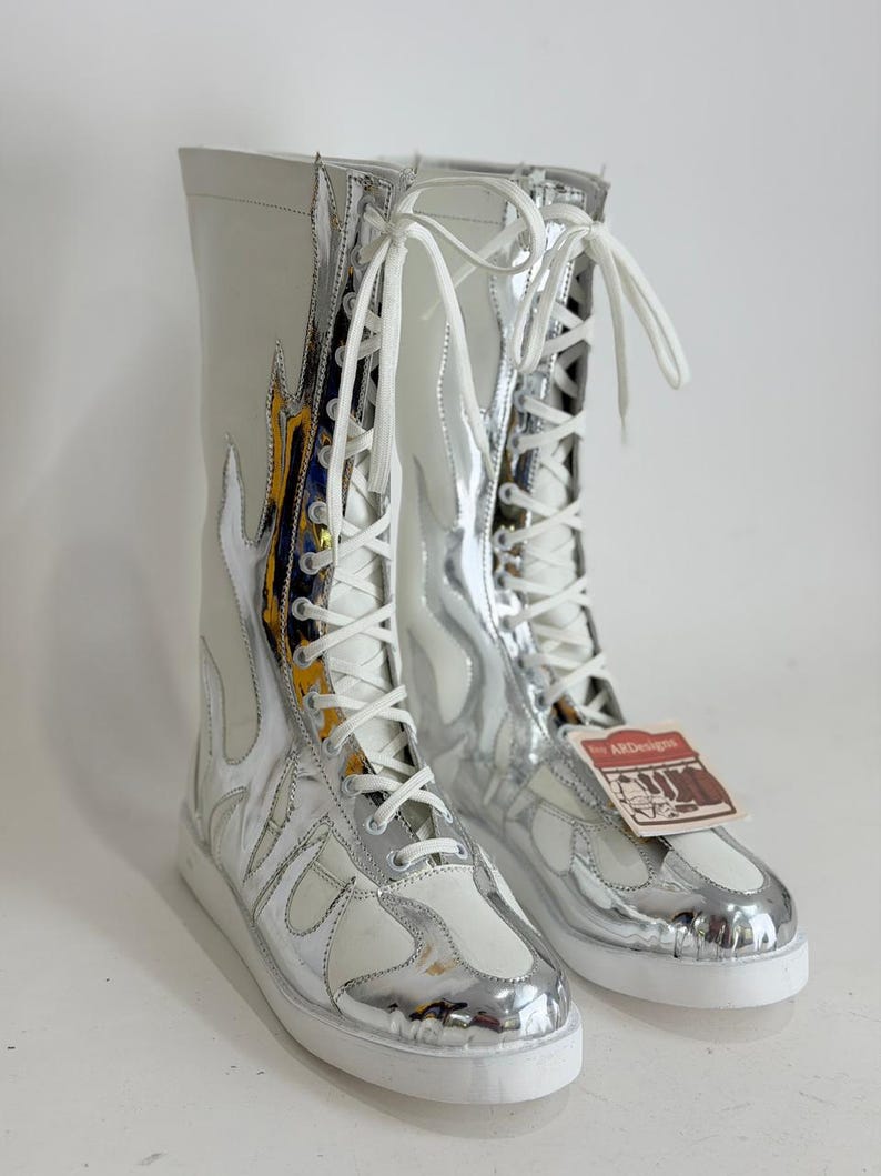 Puede incluir: Botas altas con un dise&ntilde;o de llamas blanco y plateado. Las botas tienen cordones blancos y una suela blanca. La parte superior de las botas es blanca, con detalles plateados en forma de llama. Una etiqueta est&aacute; adjunta.