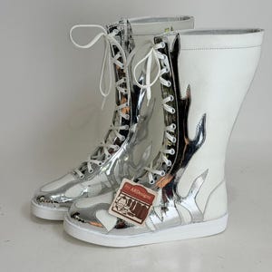 Puede incluir: Un par de botas altas blancas y plateadas con un dise&ntilde;o de llamas. Las botas tienen cordones blancos y una suela blanca. Las botas tienen una etiqueta que dice "by ARdesigns".