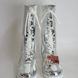 Puede incluir: Par de botas altas con un dise&ntilde;o met&aacute;lico blanco y plateado. Las botas tienen cierre de cordones y un patr&oacute;n de llamas en los laterales. Las suelas son blancas y una etiqueta con el texto "ARdesigns" est&aacute; adjunta.