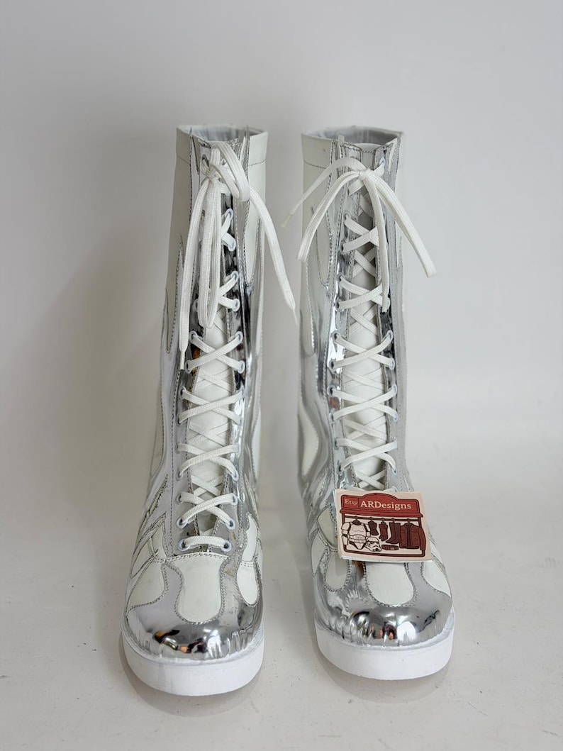 Puede incluir: Par de botas altas con un dise&ntilde;o met&aacute;lico blanco y plateado. Las botas tienen cierre de cordones y un patr&oacute;n de llamas en los laterales. Las suelas son blancas y una etiqueta con el texto "ARdesigns" est&aacute; adjunta.