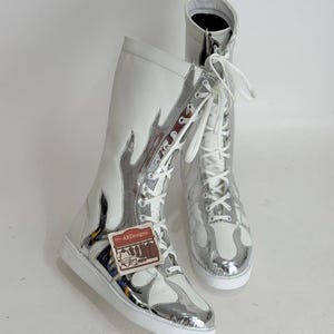 Puede incluir: Un par de botas altas blancas y plateadas con un dise&ntilde;o de llama. Las botas tienen cordones blancos y una suela blanca. Las botas tienen una etiqueta que dice "ARDesigns".