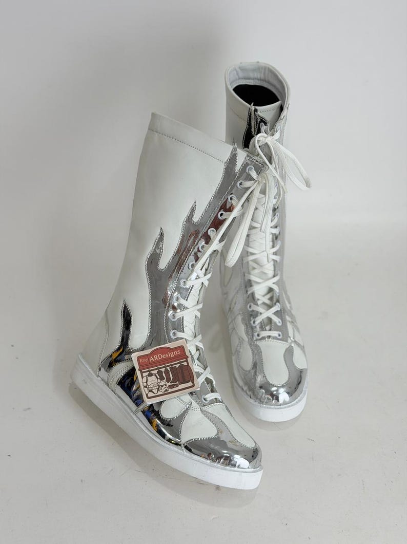 Puede incluir: Un par de botas altas blancas y plateadas con un dise&ntilde;o de llama. Las botas tienen cordones blancos y una suela blanca. Las botas tienen una etiqueta que dice "ARDesigns".