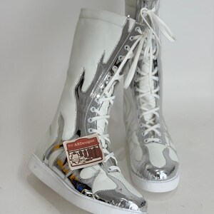 Puede incluir: Par de botas altas blancas y plateadas con dise&ntilde;o de llama. Las botas tienen cordones blancos y suela blanca. Las botas tienen una etiqueta que dice "ARDesigns".