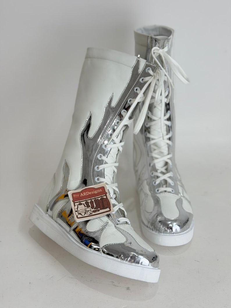 Puede incluir: Par de botas altas blancas y plateadas con dise&ntilde;o de llama. Las botas tienen cordones blancos y suela blanca. Las botas tienen una etiqueta que dice "ARDesigns".