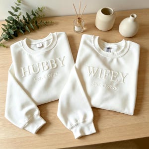 May include: Two off-white crewneck sweatshirts. One features the word "HUBBY" and the date "EST. 17.07.26" embroidered on the front. The other has the word "WIFEY" and the date "EST. 17.07.26" embroidered on the front.