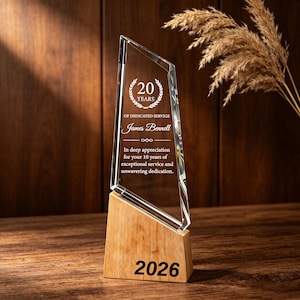 May include: A clear, angular glass award plaque mounted on a light-toned wooden base. The glass features an etched laurel wreath, the text 20 YEARS OF DEDICATED SERVIGE, the name James Bennell, and an appreciation message. The year 2026 is printed on the base.